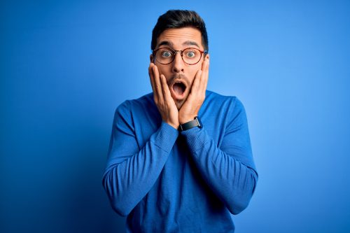 Young man with beard wearing casual blue sweater and glasses over blue background afraid and shocked