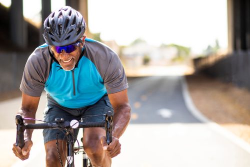 Пожилой темнокожий мужчина мчится на своем шоссейном велосипеде, готовясь к гонке.  Он улыбается и носит шлем.