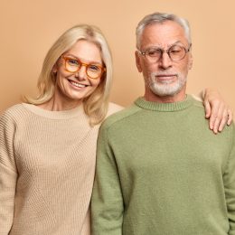 Elderly husband and wife pose for family portrait embrace smile positively dressed in eyewear jumpers stand against brown studio background.