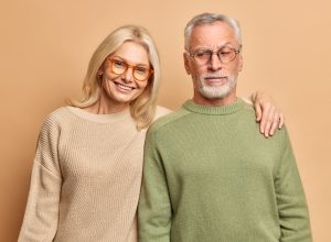 Elderly husband and wife pose for family portrait embrace smile positively dressed in eyewear jumpers stand against brown studio background.