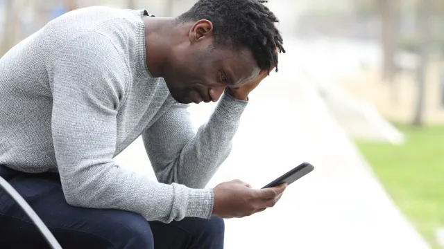 Side view portrait of a sad black man checking his cell phone while sitting on a bench in a park