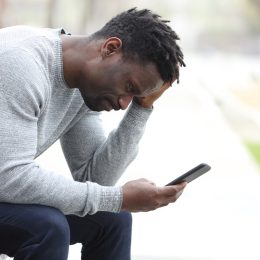 Side view portrait of a sad black man checking his cell phone while sitting on a bench in a park