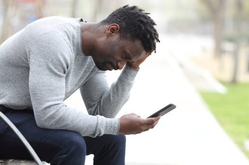 Side view portrait of a sad man checking his cell phone while sitting on a bench in a park