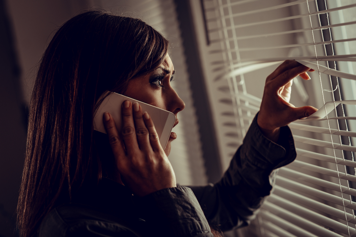 A scared young woman looking through the window blinds while on the phone.