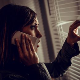 A scared young woman looking through the window blinds while on the phone.