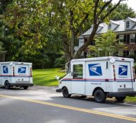 Two United States Postal trucks parked on a suburban Boston street in a quiet neighborhood.
