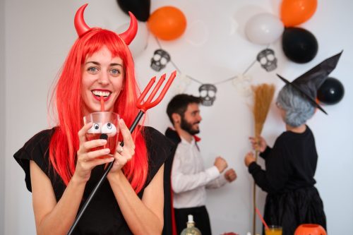 Young woman dressed as devil smiling drinking a red cocktail with eyes at costume Halloween party in a house.