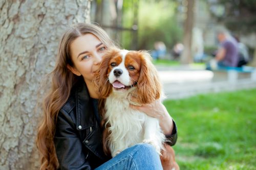 Young beautiful woman with long curly hair playing with her dog in the park .She is wearing black jacket, jeans,boots and hat .