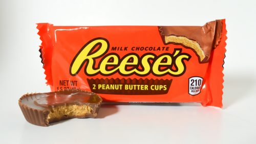 A partially eaten Reese's peanut butter cup in front of a package of peanut butter cups with a white background