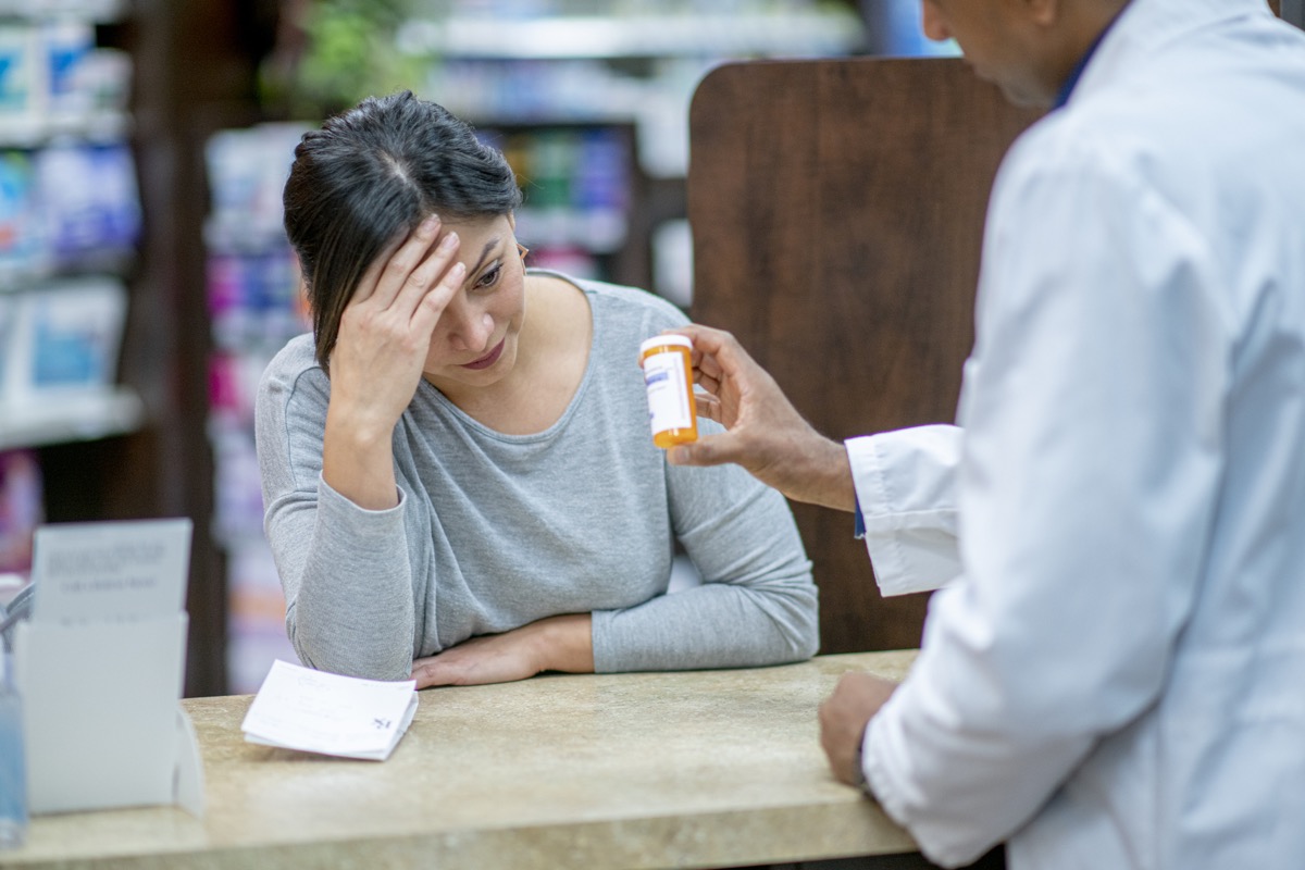 A stressed customer with her head in her hand leans on the counter and discusses medication with the pharmacist.