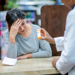 A stressed customer with her head in her hand leans on the counter and discusses medication with the pharmacist.