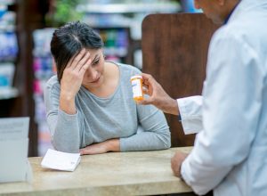 A stressed customer with her head in her hand leans on the counter and discusses medication with the pharmacist.