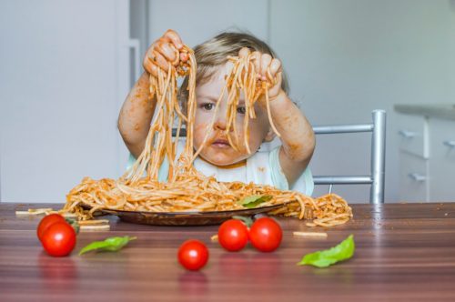 Child getting messy eating spaghetti with tomato sauce from a large plate, by itself with his hands