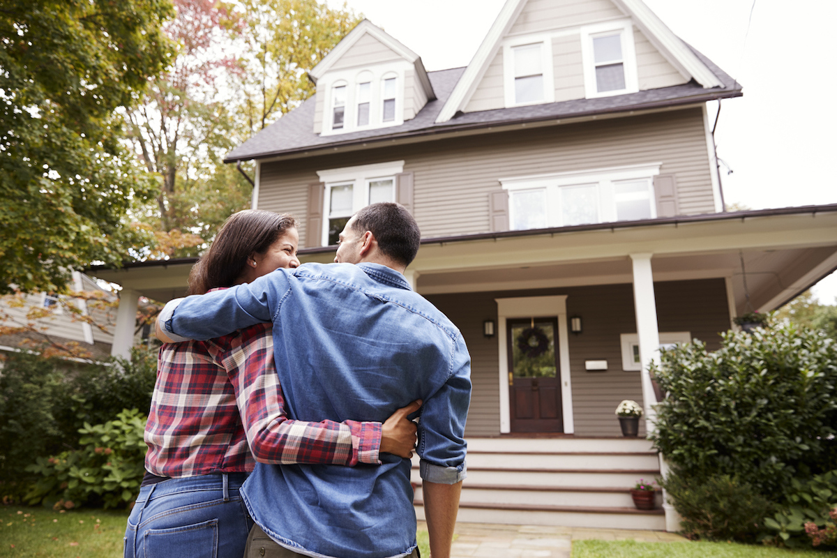 A couple hugging and looking at each other outside of a Victorian house.
