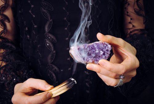 Close up view of a woman in a black lace dress, cleansing an amethyst crystal with a sage stick.