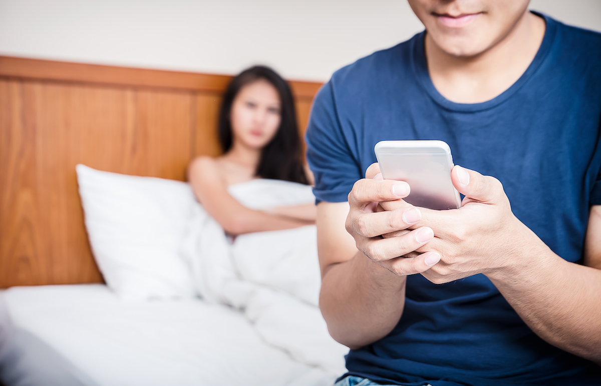 A couple in bed with the man sitting up texting while the woman looks suspicious in the background.