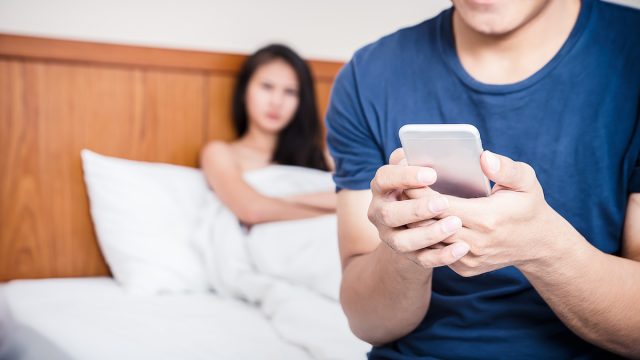 A couple in bed with the man sitting up texting while the woman looks suspicious in the background.