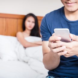 A couple in bed with the man sitting up texting while the woman looks suspicious in the background.