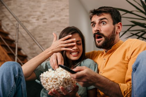 Couple watching a scary horror movie on a TV, both sitting on sofa and holding a popcorn pot. The woman is scared, man is shocked.