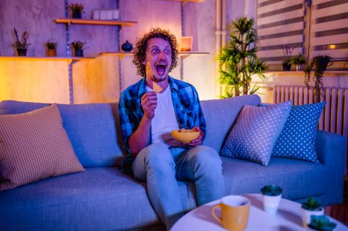 Young scared man watching horror movie and eating snacks in the living room at night