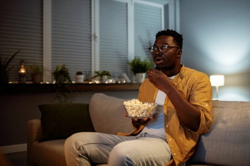 Serious young guy taking some popcorn from a bowl while watching horror movie