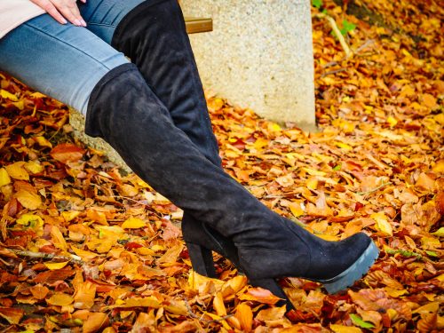 Close up of a woman's legs wearing jeans and black knee-high boots with orange leaves on the ground.
