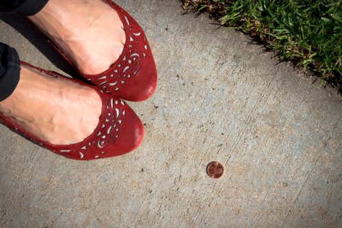 Close up of a women in red flat shoes with a lucky heads-up penny near her feet on the sidewalk.