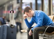 A man sitting in an airport with his luggage holding his head looking distressed
