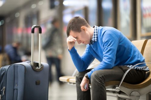 A man sitting in an airport with his luggage holding his head looking distressed