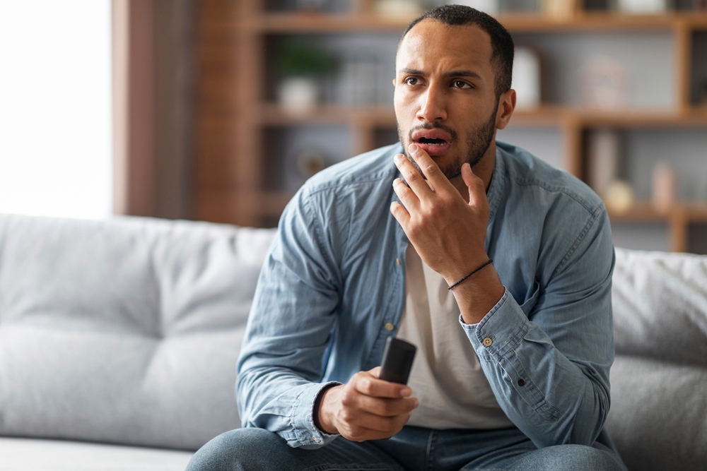 A young man holding a remote while watching TV with a confused or upset look on his face