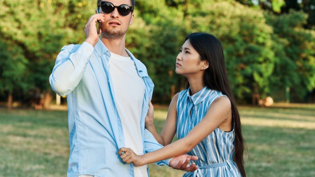 Couple arguing in a park. Man is in sunglasses on the phone not paying attention to his girlfriend who's tugging at his shirt.