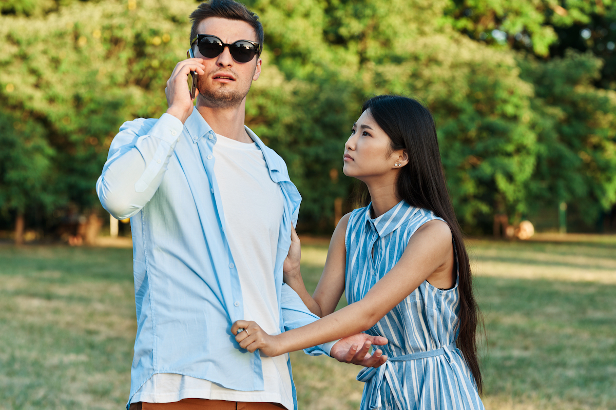 Couple arguing in a park. Man is in sunglasses on the phone not paying attention to his girlfriend who's tugging at his shirt.