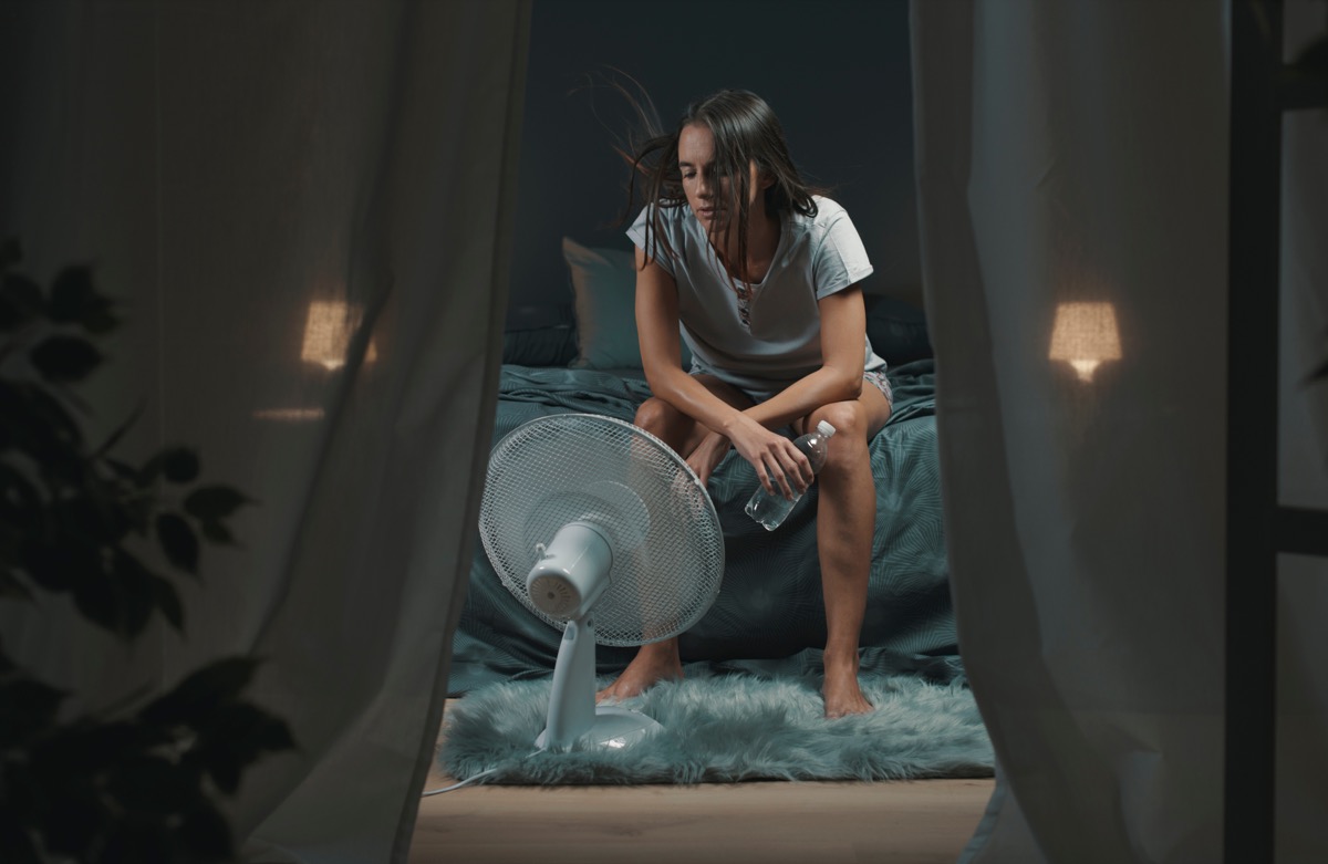 Exhausted woman suffering suring night sweats, she is holding a water bottle and sitting in front of a cooling fan in the bedroom