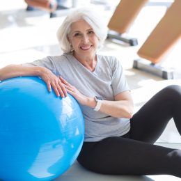An older woman with white hair sitting next to a blue yoga ball wearing black leggings and a gray t-shirt.