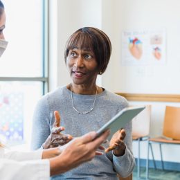 An attentive doctor listens as a female patients asks questions about recent medical test results. The doctor is holding a digital tablet which contains the test results. The doctor is wearing a protective face mask.