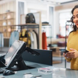 Clothing Store Checkout Cashier Counter: Woman Retail Sales Manager Accept NFC Smartphone and Credit Card Payments from a Young Female Customers for Clothes.