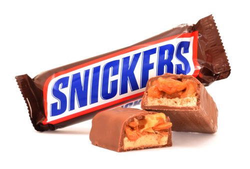 Close up of a Snickers candy bar broken in half showing chocolate bar with peanuts, caramel and nougat with the wrapper in the background.