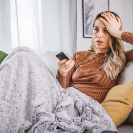 Young cheerful woman watching bed news on TV while switching channels with remote control at home.