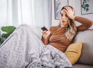 Young cheerful woman watching bed news on TV while switching channels with remote control at home.