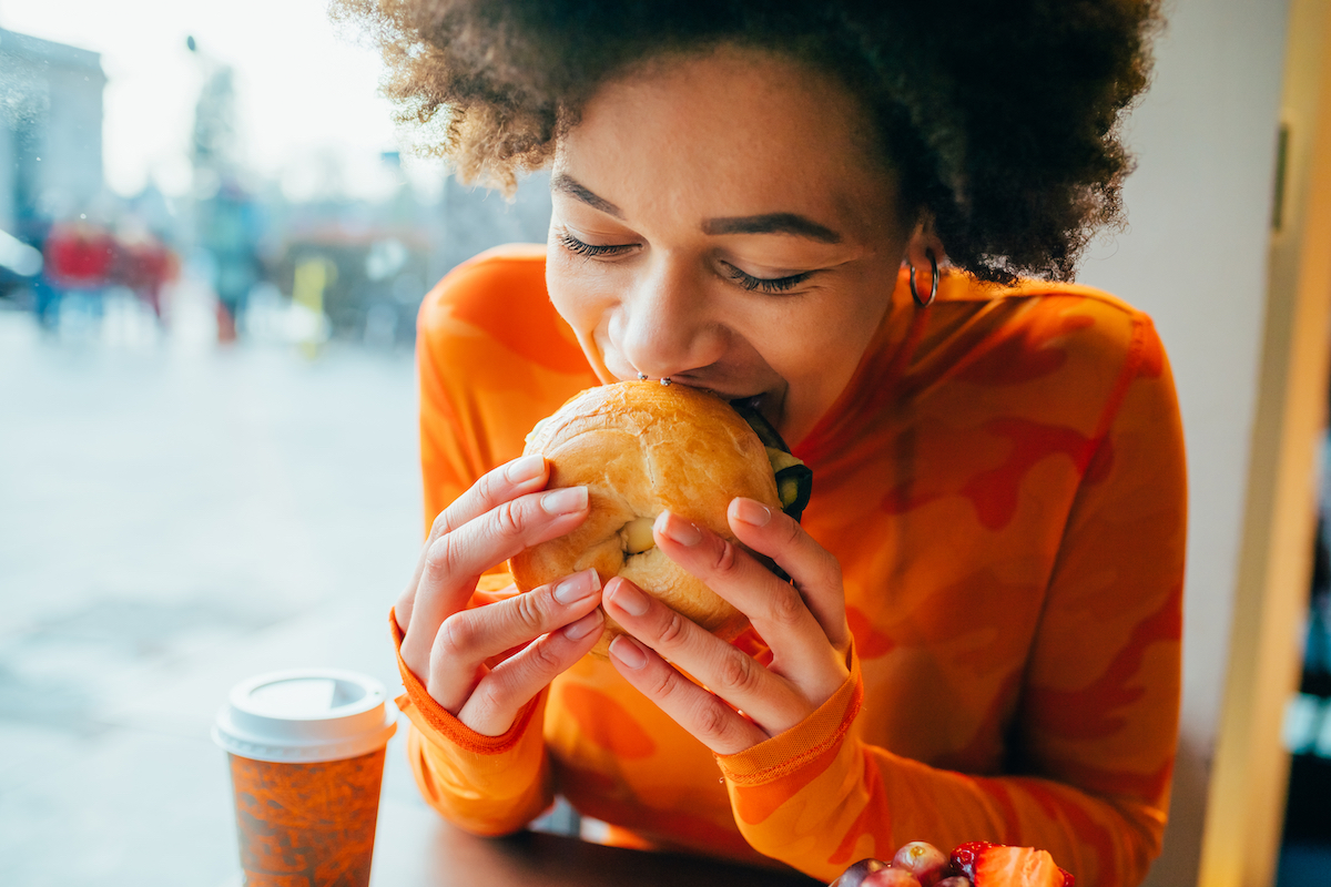 A woman sitting in a cafe eating a bagel while wearing a bright orange shirt.