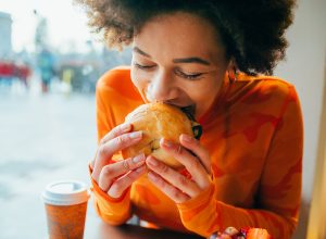 A woman sitting in a cafe eating a bagel while wearing a bright orange shirt.