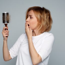 A young woman with short blonde hair looking nervously at her hair brush with a lot of hair in it.
