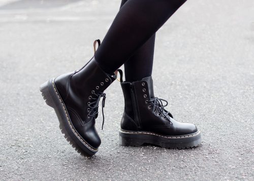 Chân nữ mặc quần tất đen và giày chiến đấu đen trên nền đường phố xám xịt.