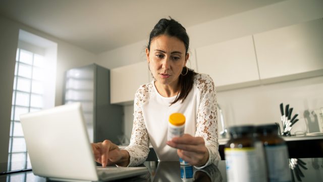 A woman sitting at her laptop and looking at a prescription pill bottle