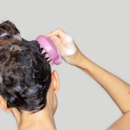 Close up of the back of a woman's head in the shower with shampoo in her hair and using a scalp massager.