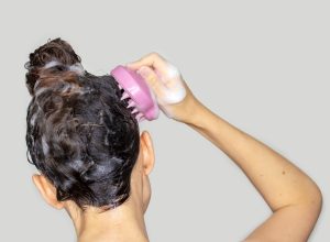 Close up of the back of a woman's head in the shower with shampoo in her hair and using a scalp massager.