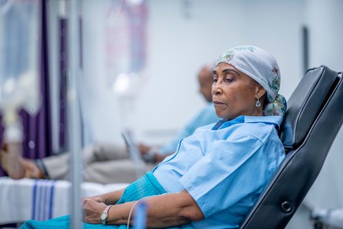 A woman wearing a head scarf lies on a hospital bed and looks to the side in contemplation. She is wearing a head scarf and a hospital gown and there is a IV drip next to her.