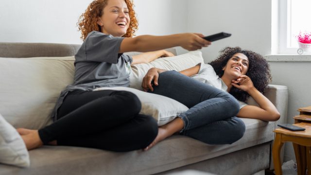 Two young women sitting on a couch using a remote and watching TV