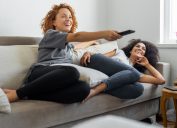 Two young women sitting on a couch using a remote and watching TV