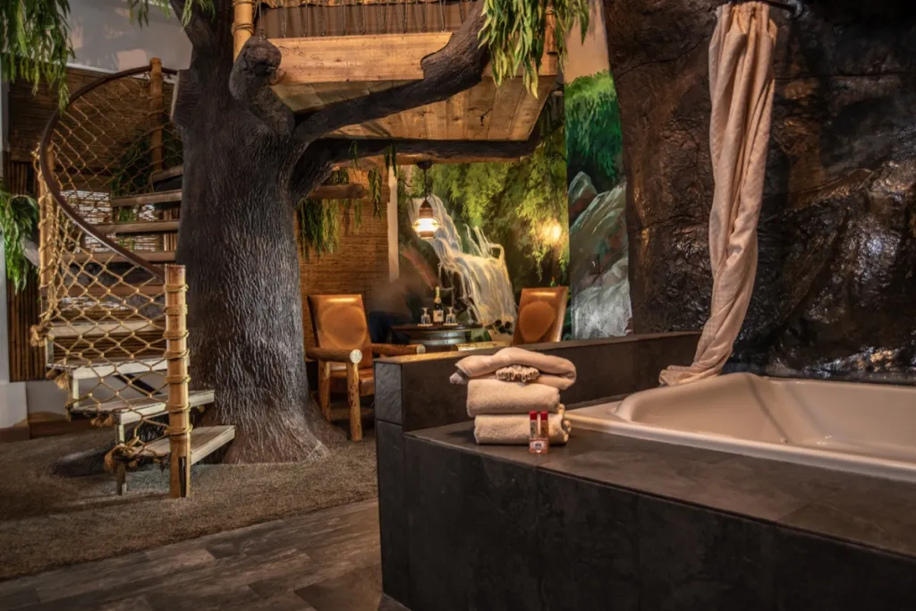 A themed hotel room with a waterfall bath, chairs, jungle wallpaper and a staircase to a treehouse bed